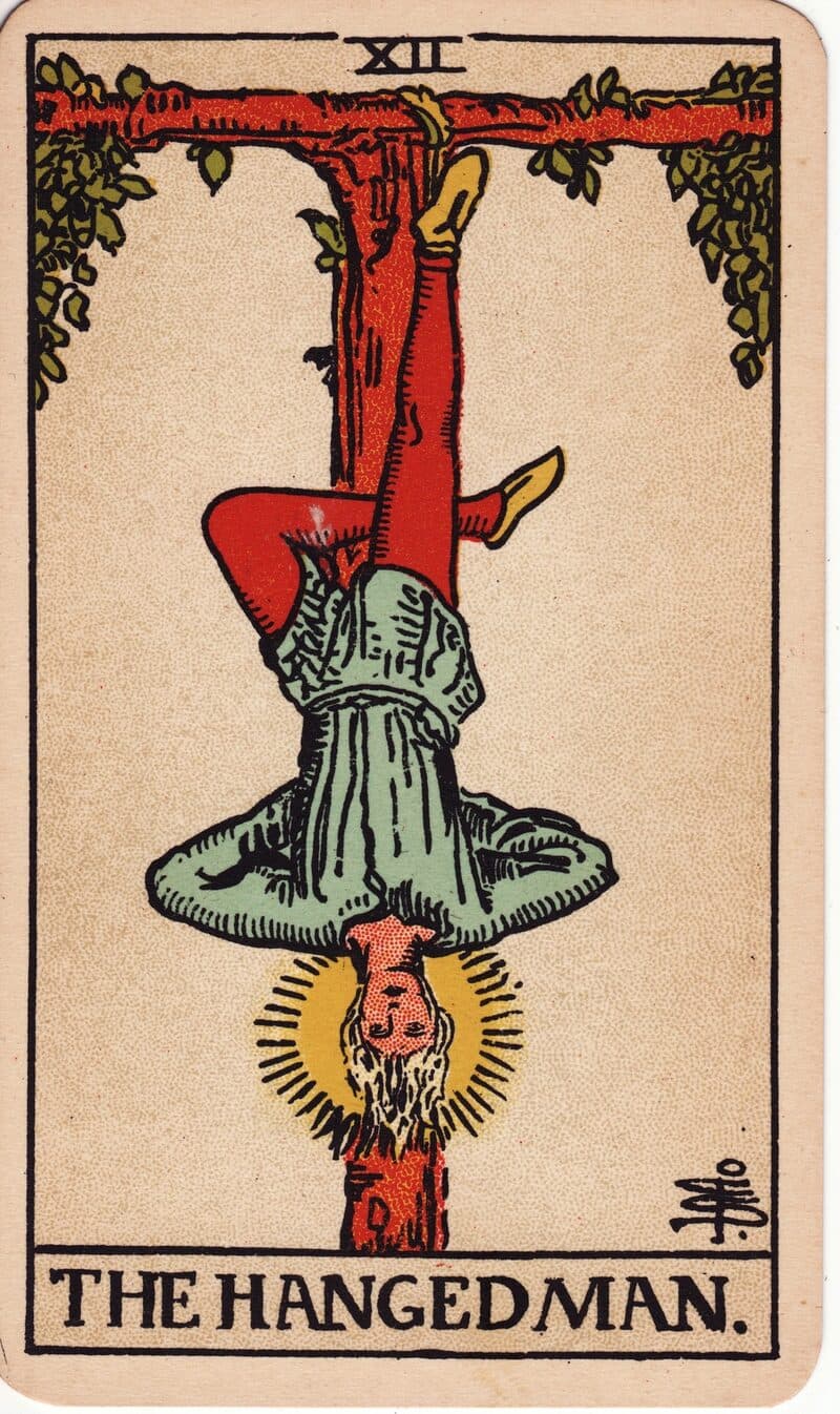 The Hanged Man tarot card — figure suspended upside-down from a living tau cross with a halo of light, Rider-Waite-Smith deck